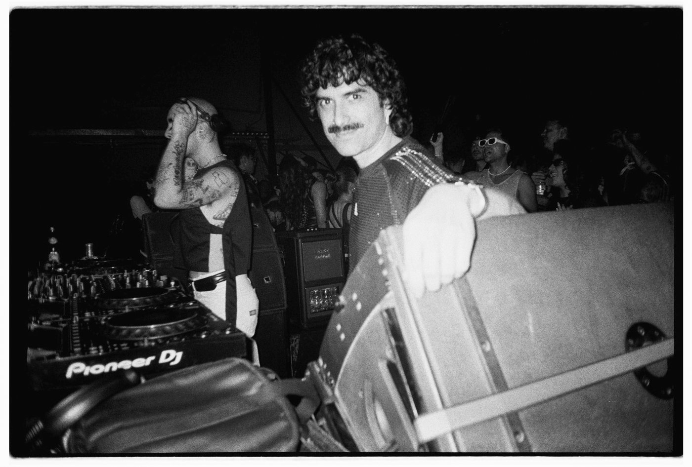 Mustached person leaning on a speaker at a DJ booth