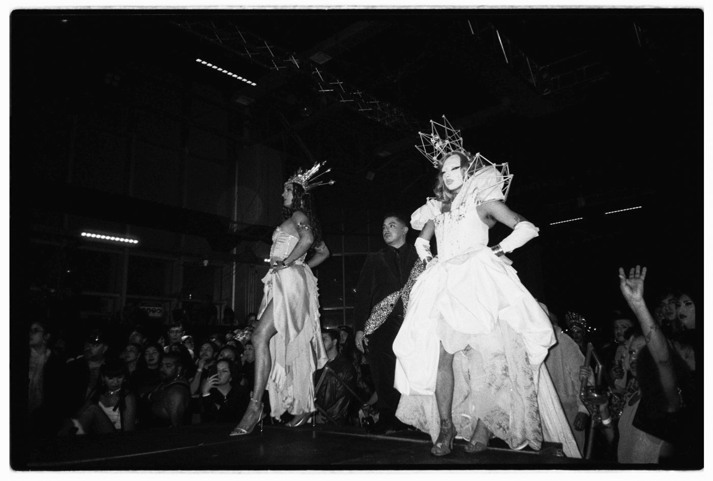 Two people in white gowns and crowns on a runway before a crowd