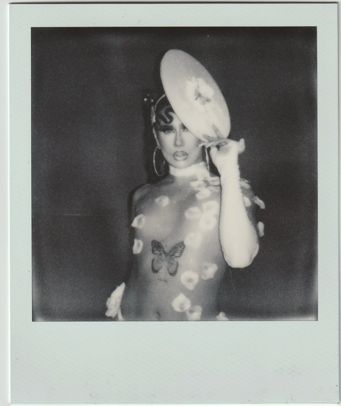Person in a sheer floral bodysuit holding a large hat to their head